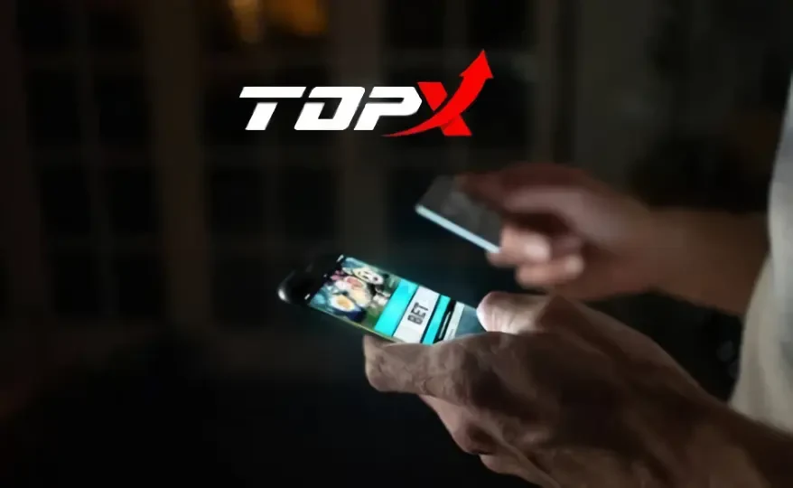 Mãos segurando um celular em ambiente escuro, com o logotipo da TopX